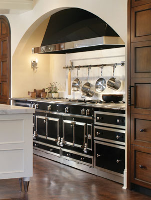 A black and polished-steel La Cornue range set beneath a plaster arch in a fitted kitchen, with copper pans hanging above it.